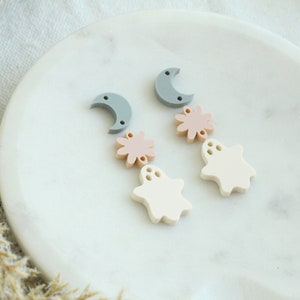 May include: A pair of dangle earrings featuring a crescent moon, a pink flower, and a white ghost charm. The earrings are made of polymer clay and have a matte finish.