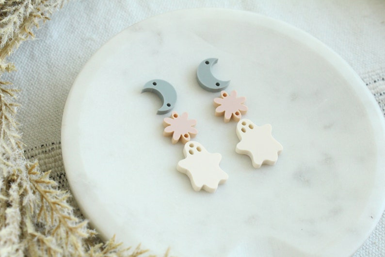 May include: A pair of dangle earrings featuring a crescent moon, a pink flower, and a white ghost charm. The earrings are made of polymer clay and have a matte finish.