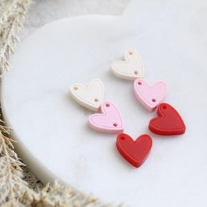 May include: A collection of heart-shaped beads in various colors: white, pink, and red. Each bead has two small holes for stringing. The beads are arranged on a white, heart-shaped surface, suitable for jewelry making or crafts.