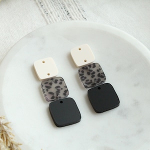May include: A pair of statement earrings with three stacked square-shaped pieces. The top piece is white, the middle piece is a black and white animal print, and the bottom piece is black.