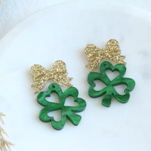May include: A pair of green shamrock earrings with gold glitter bows. The shamrocks are made of green acrylic and have a marbled effect. The bows are made of gold glitter and are attached to the top of the shamrocks.