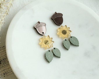 Op de afbeelding: Twee paar oorbellen met herfstthema's. Het ene paar heeft een bruine eikelbedel en een groene bladerenbedel. Het andere paar heeft een gele zonnebloembedel en een groene bladerenbedel.
