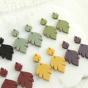 May include: A collection of leaf-shaped earrings in various colors, including black, green, yellow, and burgundy. The earrings are arranged on a white, cloud-shaped surface, with a textured white background.