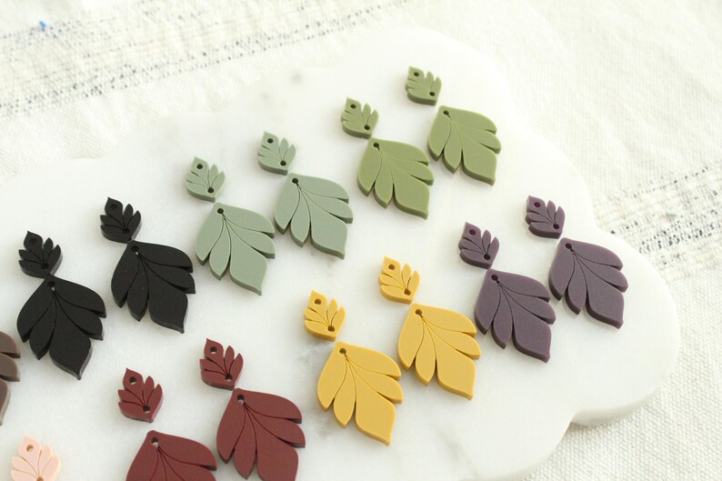 May include: A collection of leaf-shaped earrings in various colors, including black, green, yellow, and burgundy. The earrings are arranged on a white, cloud-shaped surface, with a textured white background.