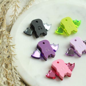 May include: Five colorful bat-shaped acrylic charms in black, green, purple, pink, and light pink. Each charm has mirrored wings. The charms are arranged on a white, round surface.