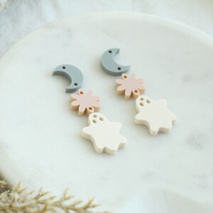 May include: A pair of dangle earrings with a moon, flower, and ghost charm. The charms are made of white, light pink, and gray acrylic.