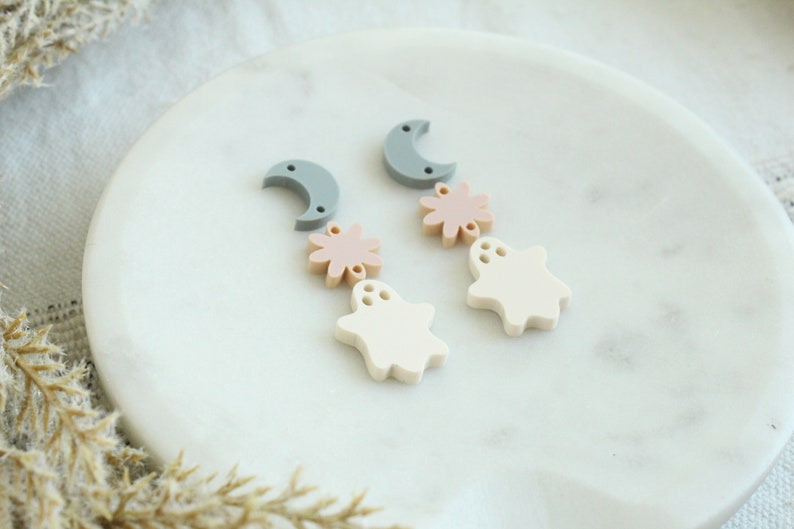 May include: A pair of dangle earrings with a moon, flower, and ghost charm. The charms are made of white, light pink, and gray acrylic.