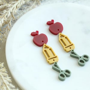 May include: A pair of school-themed earrings. Each earring features a red apple, a yellow pencil, and gray scissors. The earrings are set against a white background with a sprig of dried grass.