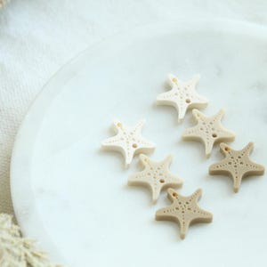 May include: Six starfish-shaped buttons in varying shades of beige and tan are arranged on a white marble surface. Each button has a detailed design and small holes for sewing, perfect for craft projects or adding a nautical touch.