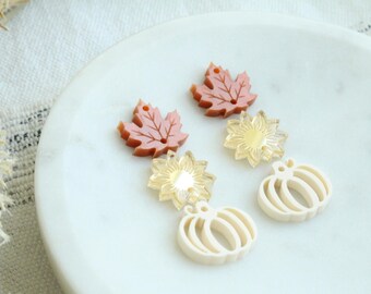May include: A collection of fall-themed earrings. The earrings feature maple leaf, sunflower, and pumpkin designs. The maple leaf earrings are a reddish-brown color. The sunflower earrings have a gold center. The pumpkin earrings are off-white.