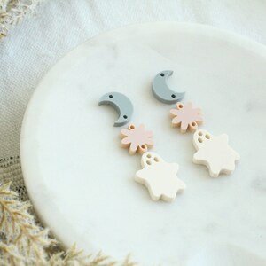 May include: A pair of dangle earrings with a crescent moon, a pink flower, and a white ghost charm. The earrings are on a white marble surface.