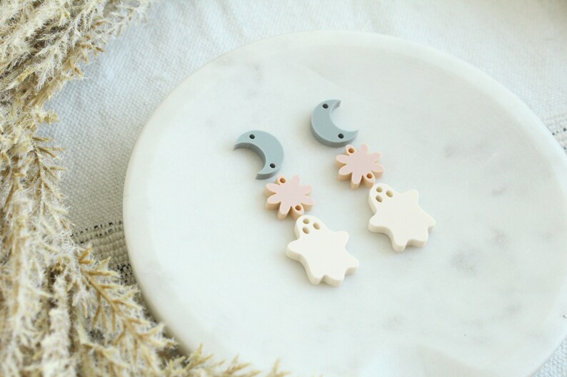 May include: A pair of dangle earrings with a crescent moon, a pink flower, and a white ghost charm. The earrings are on a white marble surface.