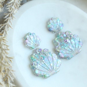 May include: Four iridescent seashell-shaped embellishments with glitter accents. The shells vary in size and are arranged on a white surface. The shells have a pearlescent sheen, reflecting shades of blue, green, and purple.