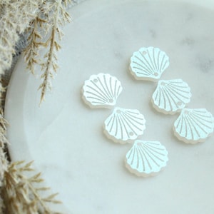May include: Six iridescent seashell-shaped craft embellishments. Each shell is white with a pearlescent sheen and detailed with etched lines. They are arranged on a white marble surface, suggesting a coastal or nautical theme.