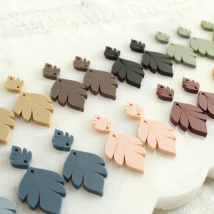 May include: A collection of leaf-shaped embellishments in various colors, including beige, brown, black, green, pink, and blue. The leaves are arranged on a white surface, showcasing their different shades and sizes. These decorative elements are ideal for crafting and DIY projects.
