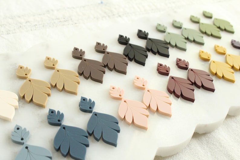 May include: A collection of leaf-shaped embellishments in various colors, including beige, brown, black, green, pink, and blue. The leaves are arranged on a white surface, showcasing their different shades and sizes. These decorative elements are ideal for crafting and DIY projects.
