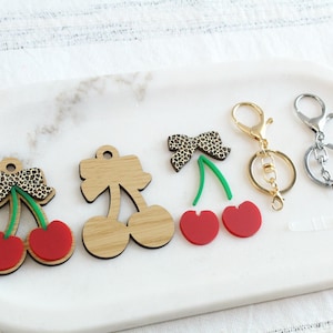 May include: A collection of cherry-themed keychains and charms. The designs include red cherries, wooden cherry shapes, and charms with leopard print bows. Two keychains have gold and silver hardware.
