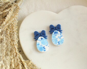 May include: Two blue and white floral egg-shaped earrings with dark blue bows on top. The earrings are set on a white, heart-shaped surface. Dried wheat stalks are in the background.