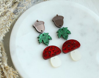 May include: Two pairs of earrings featuring autumnal themes. One pair has a red and white mushroom charm, the other has a brown acorn and a green maple leaf charm.