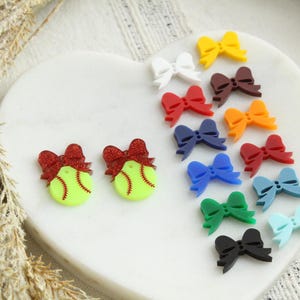 May include: A collection of colorful bow-shaped charms and two softball-themed charms with red glitter bows. The bows are in various colors, including white, red, blue, green, and yellow. The charms are on a white heart-shaped surface.
