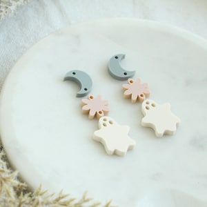 May include: A pair of earrings featuring a crescent moon, a flower, and a ghost charm in shades of gray, pink, and white.