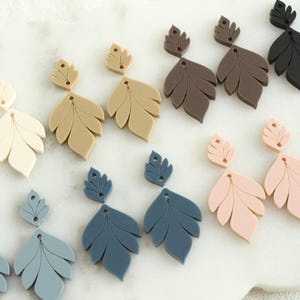 May include: A collection of leaf-shaped earrings in various colors, including cream, tan, brown, black, pink, and blue. The earrings are arranged on a white surface, showcasing their intricate design and color variations. These earrings are a stylish accessory.