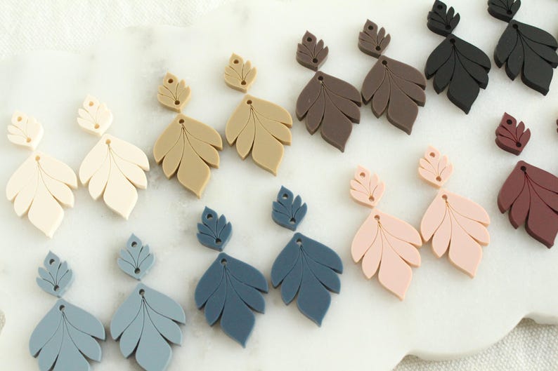 May include: A collection of leaf-shaped earrings in various colors, including cream, tan, brown, black, pink, and blue. The earrings are arranged on a white surface, showcasing their intricate design and color variations. These earrings are a stylish accessory.