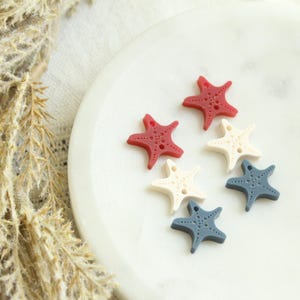 May include: Six starfish-shaped buttons in red, off-white, and blue hues. The buttons are arranged on a white, marble-like surface. The buttons have small holes for sewing and are a nautical theme.