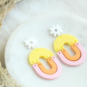 May include: A pair of colorful earrings with a yellow, orange, and pink layered arch design. Each earring is topped with a white flower charm.