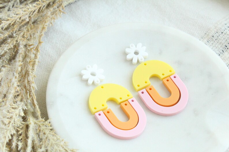 May include: A pair of colorful earrings with a yellow, orange, and pink layered arch design. Each earring is topped with a white flower charm.