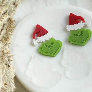 May include: Two green Grinch character acrylic blanks with red and white Santa hats. The blanks are on a white surface with two clear acrylic blanks.