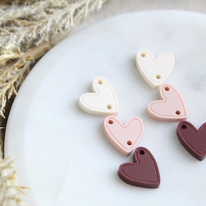 May include: Close-up of several heart-shaped beads in shades of cream, pink, and burgundy. Each bead has two small holes. The beads are arranged on a white, marble-like surface, with dried plants in the background.