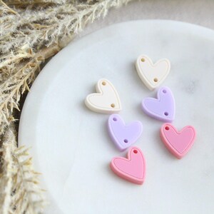 May include: A collection of heart-shaped beads in pastel colors: cream, lavender, and pink. Each bead has two small holes for stringing. The beads are arranged on a white, marble-like surface, with dried plants in the background.