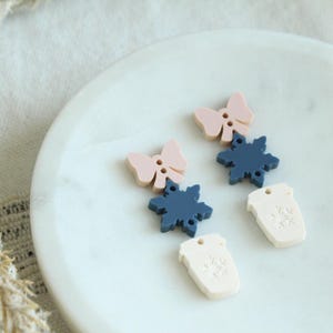 May include: A pair of dangle earrings with a pink bow, a blue snowflake, and a white coffee cup charm. The earrings are on a white marble surface.