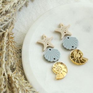 May include: A collection of decorative beach-themed embellishments. The assortment includes tan starfish, light blue scallop shells, and gold-colored nautilus shells. These small, detailed pieces are arranged on a white, marble-like surface.