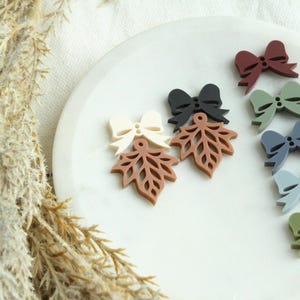 May include: A collection of colorful resin embellishments, including leaf shapes and bow shapes. The leaves are in shades of brown and the bows are in shades of red, green, blue, and black. The embellishments are arranged on a white plate.