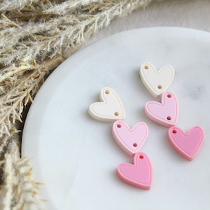 May include: A collection of heart-shaped beads in shades of pink and cream, each with two small holes for stringing. The beads are arranged on a white, marble-like surface, with dried plants in the background.