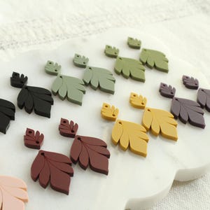 May include: A collection of leaf-shaped embellishments in various colors, including black, brown, green, yellow, and purple. The leaves are arranged on a white, cloud-shaped surface, suggesting a craft or decorative theme.