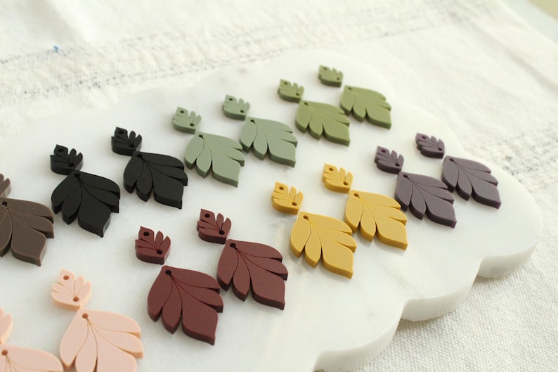 May include: A collection of leaf-shaped embellishments in various colors, including black, brown, green, yellow, and purple. The leaves are arranged on a white, cloud-shaped surface, suggesting a craft or decorative theme.