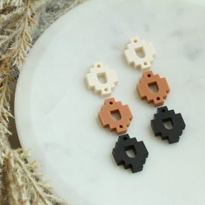 May include: A collection of six geometric earrings in a pixelated design. The earrings are in three colors: off-white, terracotta, and black. They are arranged on a white, marble-like surface.