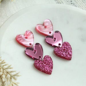 May include: Three pink heart-shaped charms with different finishes. The top charm is a translucent pink with a swirl pattern. The middle charm is a solid pink with a reflective finish. The bottom charm is a pink glitter heart.