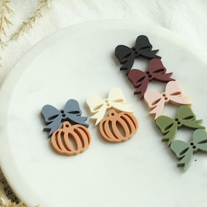 May include: A collection of colorful pumpkin and bow-shaped embellishments in various shades of blue, brown, cream, pink, green, and black. The decorative items are arranged on a white plate, perfect for fall-themed crafts and decorations.