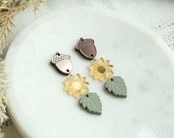 May include: Two pairs of earrings featuring autumnal themes. Each pair includes a brown acorn charm, a yellow sunflower charm, and a green leaf charm.