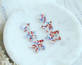 Op de afbeelding: Transparante stervormige versieringen gevuld met rode, witte en blauwe glitter. De sterren zijn verspreid over een wit marmeren oppervlak. Deze decoratieve sterren zijn ideaal voor knutselen en patriottische projecten.