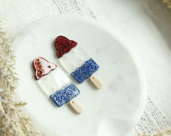 Op de afbeelding: Twee rode, witte en blauwe glitter ijslolly-charms. De charms zijn gevormd als ijslolly's met een rood, wit en blauw glitterontwerp. De charms bevinden zich op een wit oppervlak.