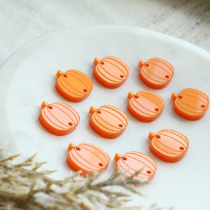 May include: A collection of orange pumpkin-shaped beads with a stem and two small holes, arranged on a white, oval-shaped dish. The beads have a striped design and are likely used for crafting or jewelry making.