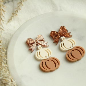 May include: Two sets of fall-themed earrings. Each set features a glittery bow, a cream-colored pumpkin, and a brown pumpkin. The earrings are arranged on a white, circular surface, with dried plants in the background.