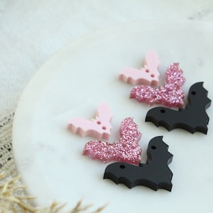 May include: Three bat-shaped buttons, two are pink with glitter and one is black. The buttons have a hole in the center for sewing.