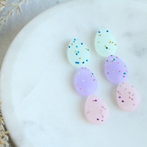 May include: Five pastel-colored egg-shaped earrings with glitter accents. The earrings are arranged in a row on a white marble surface.