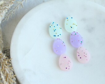 May include: Five pastel-colored egg-shaped earrings with glitter accents. The earrings are arranged in a row on a white marble surface.